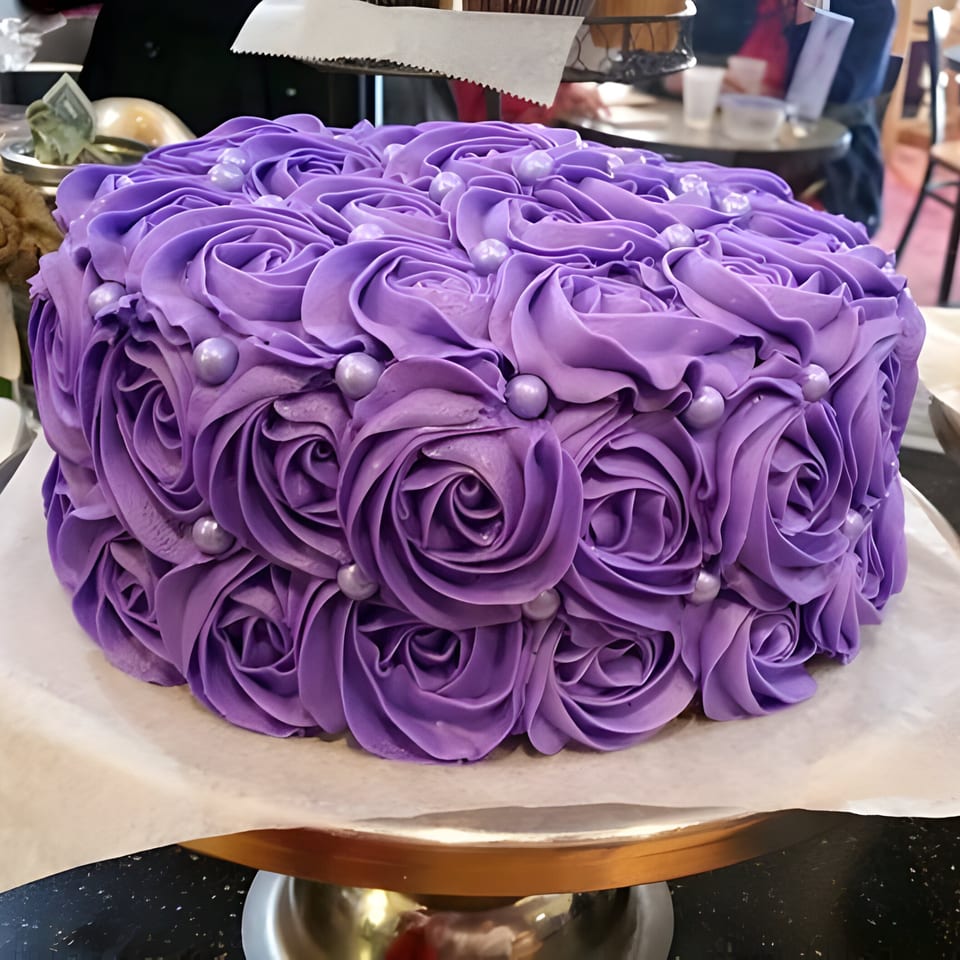 A vibrant purple cake with intricate rose-shaped frosting and small pearl decorations.