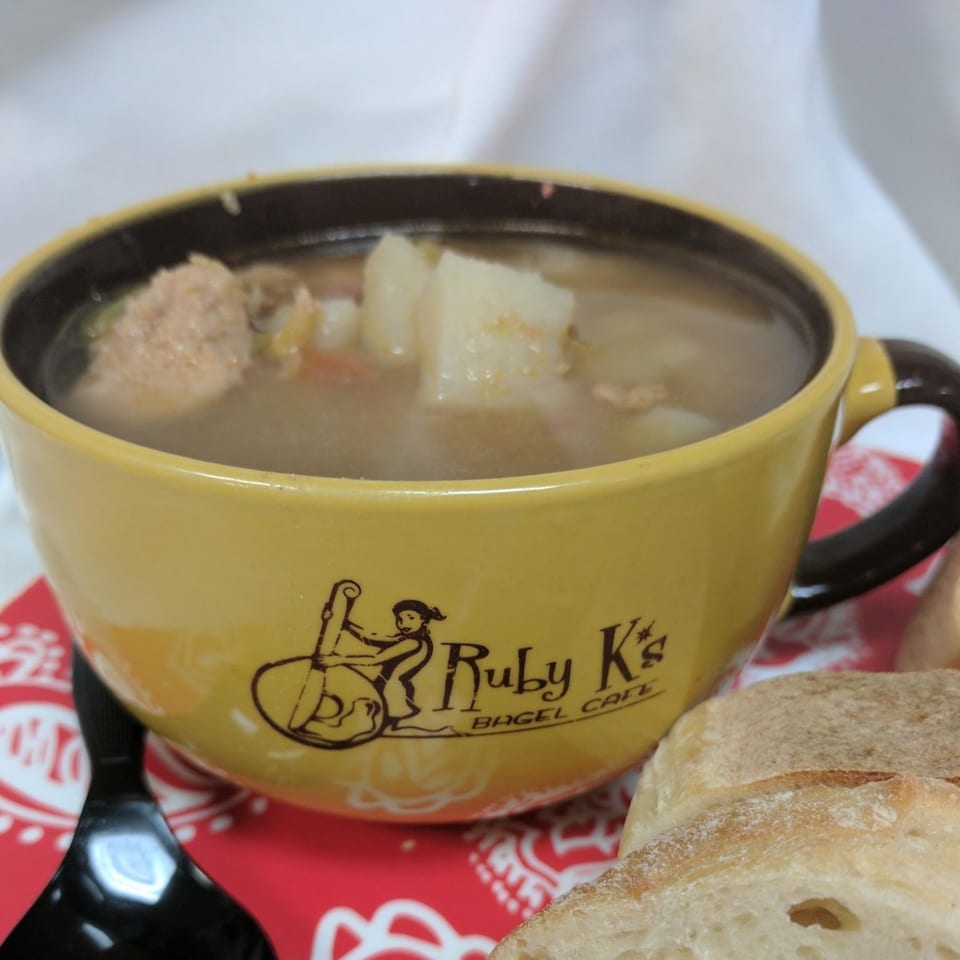 A yellow Ruby K's Bagel Cafe mug filled with soup, next to a slice of bread.